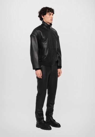 Black leather jacket with high collar, zip front, and wide shoulders, paired with matching black leather pants and lace-up boots.