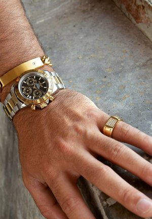 Luxuriöse Chronographenuhr mit schwarzem Zifferblatt, goldenen Akzenten und einem Silber-/Metallarmband; goldenes Manschettenarmband und rechteckiger Ring mit diamantähnlichen Details.