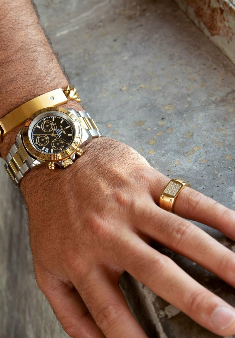 Luxuriöse Chronographenuhr mit schwarzem Zifferblatt, goldenen Akzenten und einem Silber-/Metallarmband; goldenes Manschettenarmband und rechteckiger Ring mit diamantähnlichen Details.