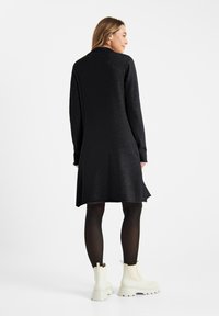 Zwart lang mouwend jurk met een ontspannen pasvorm, gestructureerde stof en een licht uitlopende zoom, gecombineerd met witte enkellaarsjes en zwarte maillots.