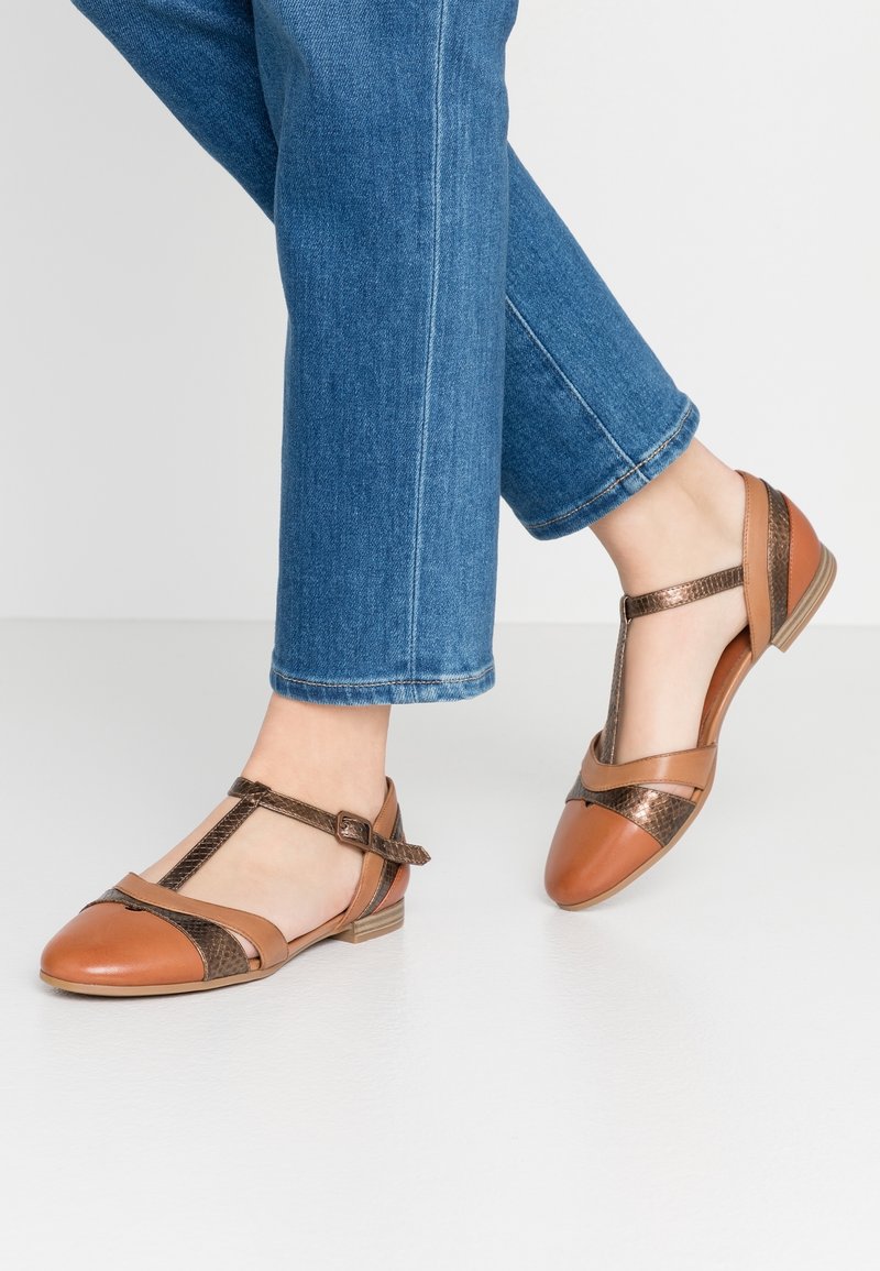 Femme portant des ballerines en cuir marron à bride en T avec des accents métalliques, associées à un jean bleu court, debout sur un sol blanc.