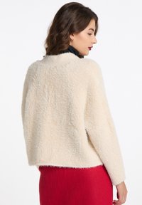 Lys cremefarvet sweater med en løs pasform, brede ærmer og en blød tekstur. Har en rund halsudskæring og bæres over en rød nederdel.