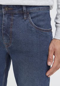 Tamnoplave traperice s kontrastnim narančastim šavovima, dizajn s pet džepova i glatkom teksturom, prikazane s rukom koja počiva na kuku.