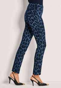 Vrouw in slanke blauwe panterprint broek en zwarte hoge slingback schoenen met gouden teentips, zijwaarts staand tegen een lichte muur.