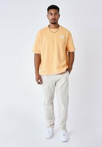 T-shirt arancione a maniche corte in maglia con una finitura testurizzata e un'etichetta a taschino, abbinato a pantaloni beige chiaro e sneakers bianche.