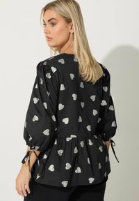 Blusa nera con motivi a cuore argentati, maniche a sbuffo con lacci e orlo peplum. Tessuto liscio con un design giocoso e audace.
