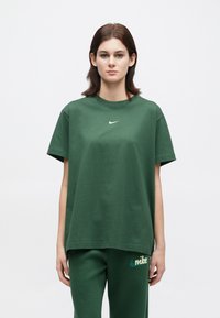 Νεαρός στέκεται μπροστά φορώντας σκοτεινό πράσινο μπλουζάκι Nike και ταιριαστούς φόρμες κατά ενός λευκού φόντου.