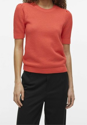 Kobieta ubrana w koralowy, krótki sweter o teksturowanym splocie, w połączeniu z czarnymi spodniami, stojąca na tle jednolitego białego tła.