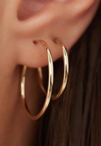 Boucles d'oreilles en or en forme de double anneau avec une surface lisse et réfléchissante, suspendues au lobe de l'oreille, sur fond de cheveux sombres.