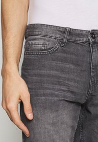 Jeans de denim gris con una sutil textura desgastada, un bolsillo delantero y un cierre de botón. Vista en primer plano de la cinturilla con una mano descansando sobre ella.