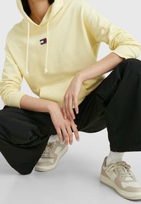 Sudadera amarilla con un bolsillo frontal, cordones ajustables y un parche con logo. Combinada con pantalones negros holgados y zapatillas beige.