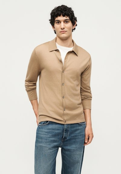 Νέος άνδρας με σγουρά μαλλιά που φοράει μπεζ ζακέτα με κουμπιά πάνω από λευκό πουκάμισο και μπλε τζιν, στέκεται μπροστά σε απλό ανοιχτό φόντο.