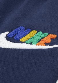 Mørkeblå plagg med en teksturert, flerfarget brodert logo i gult, grønt, oransje og blå detaljer ved siden av en hvit swoosh.