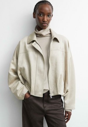 Frau mit geflochtenem Haar, die eine beige Wildlederjacke über einem hellen Rollkragenpullover und dunklen Hosen vor einem einfarbig weißen Hintergrund trägt.