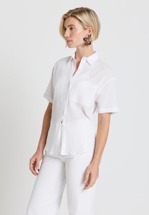 Femme blonde portant une chemise blanche oversize boutonnée, un pantalon blanc, de grandes boucles d'oreilles florales foncées et des bagues en or, debout devant un fond uni.