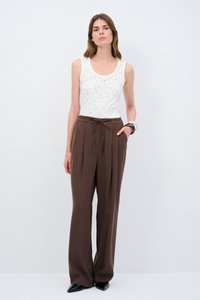 Witte mouwloze top met decoratieve zilveren studs; losse bruine broek met een verstelbare taille; zwarte puntige schoenen.