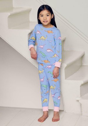 Ragazza giovane in piedi scalza accanto a scale bianche, che indossa un pigiama blu con stampe colorate di dinosauri e polsini rosa.