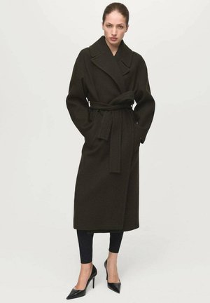 Długi, ciemnozielony wełniany płaszcz z paskiem w talii, dużym kołnierzem i bocznymi kieszeniami, zestawiony z czarnymi dopasowanymi spodniami i szpiczastymi szpilkami.