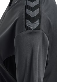 Mörkgrå atletisk topp med en slät textur. Har en kontrasterande svart panel med silverfärgade chevron-accenter längs axeln.