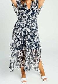 Kvinna iklädd en svart och vit blommig midiklänning i genomskinligt tyg, stående med höjda armar, iförd vita sandaler med öppen tå och klack.