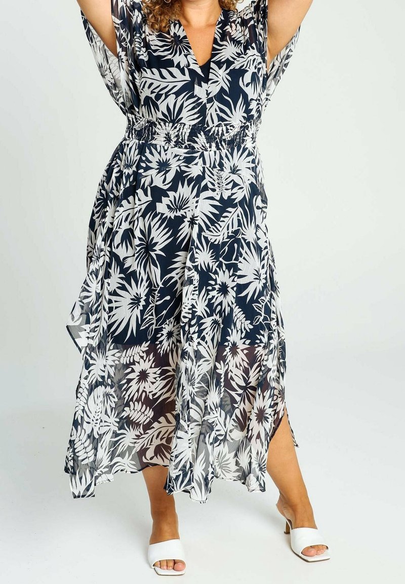 Kvinna iklädd en svart och vit blommig midiklänning i genomskinligt tyg, stående med höjda armar, iförd vita sandaler med öppen tå och klack.