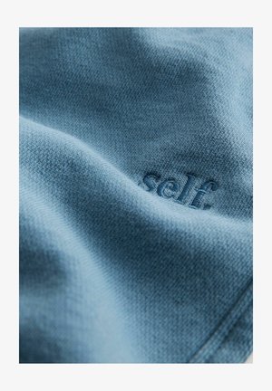Tessuto di maglia blu con un dettaglio in rilievo della scritta "self.", che mette in evidenza una texture morbida e un motivo sottile.