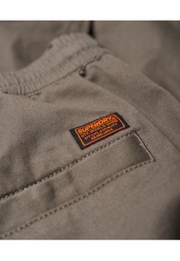 Närbild av beige tyg med sydd ficka och en liten rektangulär orange och svart Superdry-märkesetikett.