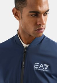 Giacca bomber navy con chiusura a zip, colletto a coste e logo ricamato EA7 Emporio Armani. Tessuto liscio con finitura opaca.