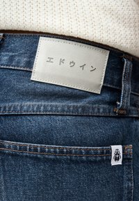 White leather label with Japanese text sewn on blue denim jeans worn with a cream knit top, showing back pocket and belt loop.