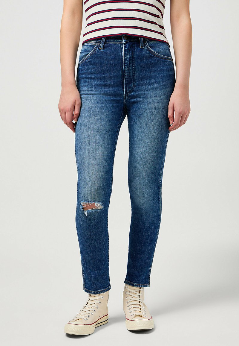 Skinny blue denim jeans with a distressed knee detail, five-pocket design, and a classic waistband. Paired with white high-top sneakers.
