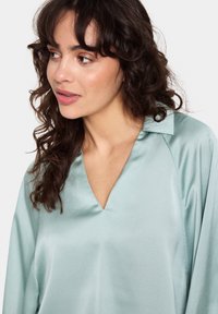 Top in satin verde menta con scollo a V e colletto, caratterizzato da una morbida texture e una vestibilità casual, cuciture a vista e maniche fluide.
