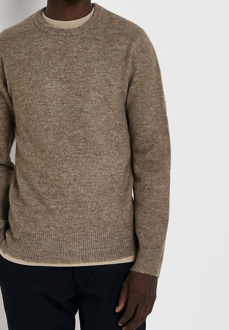 Homme portant un pull en maille col rond marron par-dessus une chemise beige, associé à un pantalon noir, debout devant un fond clair uni.