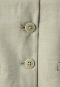 Hellbeige Stoff mit zwei großen, runden Knöpfen. Die Knöpfe sind beige und haben vier Löcher, um die herum feine Nähte sichtbar sind.