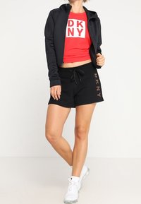 Czerwony krótki t-shirt z logo "DKNY", czarna kurtka na zamek, czarne szorty z detalem logo oraz białe buty sportowe. Gładki materiał, casualowy design.