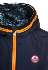 Marinblå vindjacka med en orange dragkedja och krage. Inuti har den ett texturerat blått foder. Inkluderar en rund logopatch på framsidan.