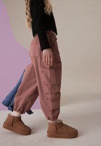 Šviesiai rožinės spalvos kargo kelnes su elastinėmis rankovėmis, turinčiomis kelis kišenes; derinamos su smėlio spalvos kulkšnių botais ir juodu ilgomis rankovėmis viršumi.