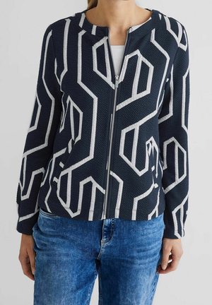 Femme portant une veste bleu marine avec des motifs géométriques blancs, fermeture éclair sur le devant, associée à un jean bleu et une chemise blanche en dessous.
