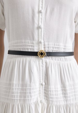 Černý kožený pásek se zlatou a černou květinovou sponou, nošený přes bílé, knoflíkové, plisované šaty. Texturovaná látka vytváří vizuální kontrast.