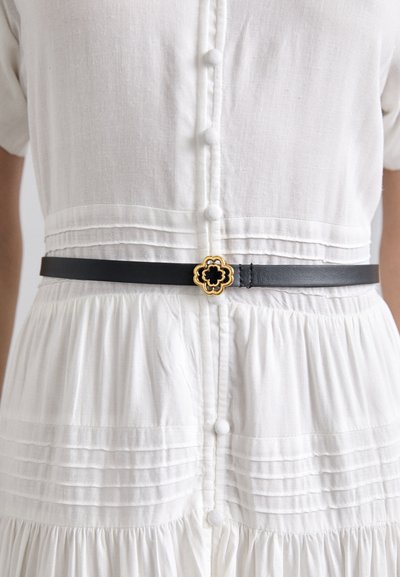 Cinturón de cuero negro con una hebilla floral en oro y negro, usado sobre un vestido blanco de botones y plisado. La tela texturizada crea un contraste visual.