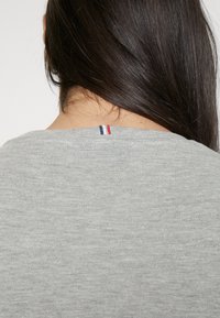Tessuto grigio di una camicia con scollo rotondo che presenta una sottile trama. I dettagli notevoli includono un piccolo accento cucito blu, bianco e rosso sul retro.