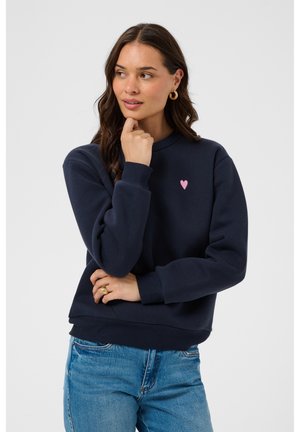 Femme aux longs cheveux bruns porte un sweat bleu marine avec un petit cœur rose sur la poitrine et un jean bleu, posant la main près du menton.