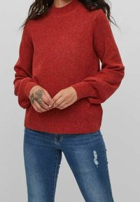 Czerwony dziergany sweter z okrągłym dekoltem i długimi bufiastymi rękawami, z prążkowanymi mankietami i dołem. Połączony z niebieskimi, przetartymi jeansami.