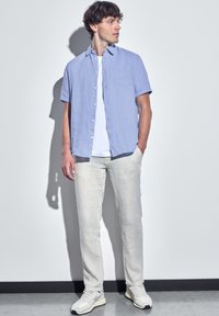 Camicetta azzurra a maniche corte con taschino sul petto, abbinata a pantaloni beige chiaro e sneakers grigie, su uno sfondo semplice.