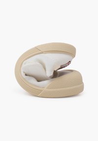 Zapato beige de tipo slip-on con suela de goma texturizada, hecho de tela suave. El zapato está doblado, mostrando su interior acolchado y los detalles de diseño.