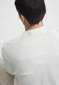 Sudadera blanca con cuello redondo, que presenta un cuello acanalado y detalles de costura en la parte posterior. Textura suave, diseño minimalista.