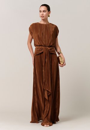 Donna che indossa un abito lungo in rame plissettato con cintura larga, tiene in mano una pochette oro metallizzato e porta orecchini a cerchio dorati su sfondo neutro.