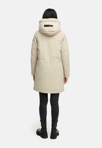 Beige lange pufferjas met een capuchon, met een zwart accent op de achterkant, gezien vanuit de achterkant. Gecombineerd met zwarte skinny jeans en laarzen.
