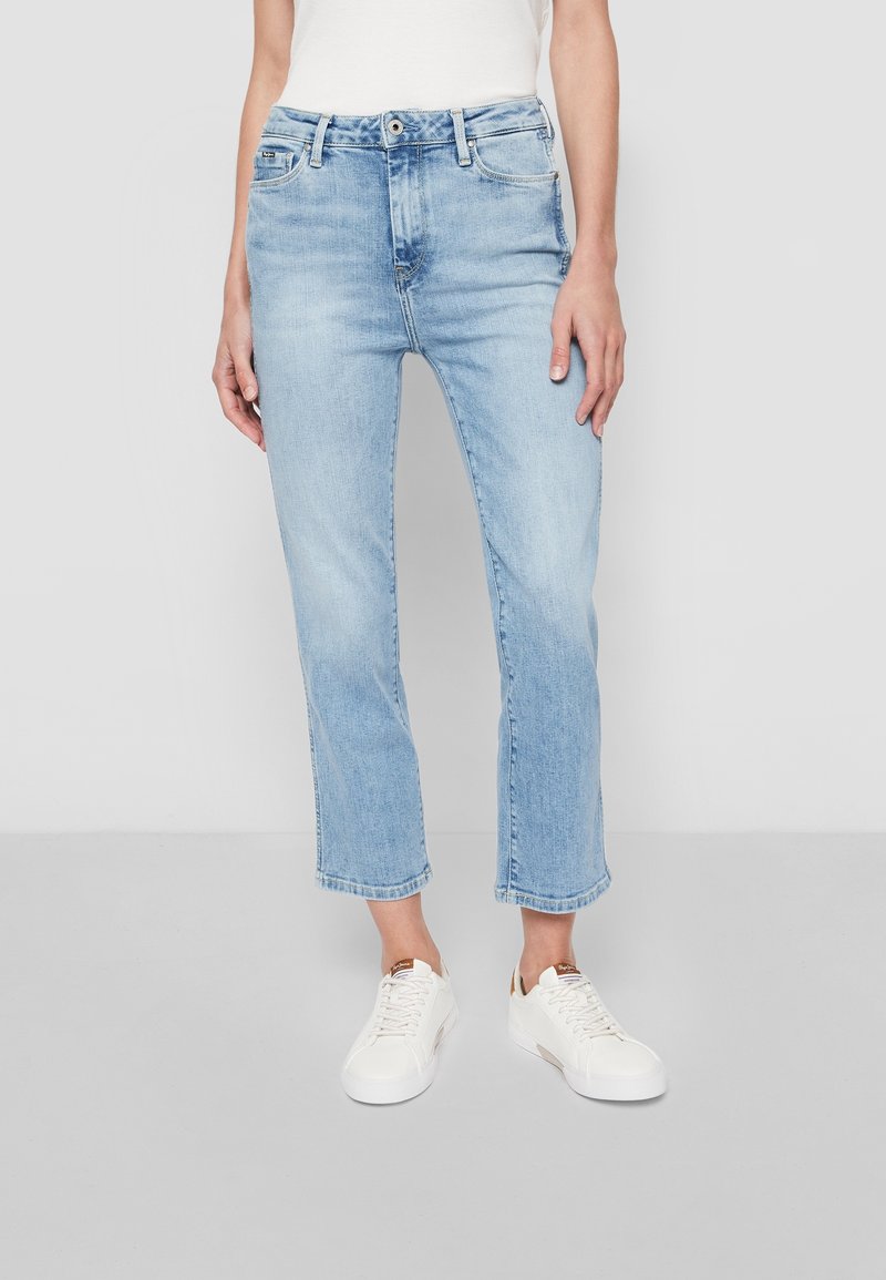 Personne portant un jean droit bleu clair raccourci, des baskets blanches et un haut blanc, debout sur un sol gris devant un fond uni.