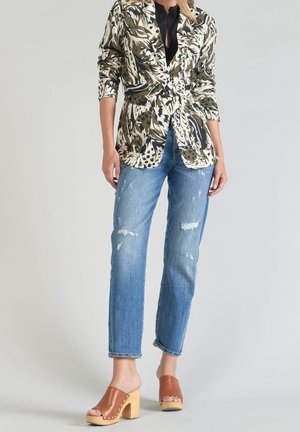 Kvinde iført en mønstret blazer, sort bluse, slidte blå jeans og brune træsko med åbne tæer og træhæl, stående mod en ensfarvet baggrund.