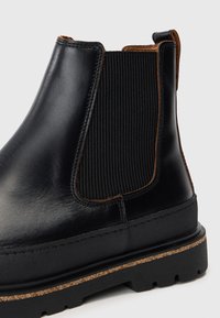 Bottine Chelsea en cuir noir avec panneau élastique sur le côté, bordure marron, languette à l'arrière et semelle en caoutchouc épaisse avec détails de crans.